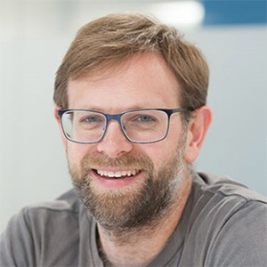 A man with fair hair, full beard, and glasses smiles.