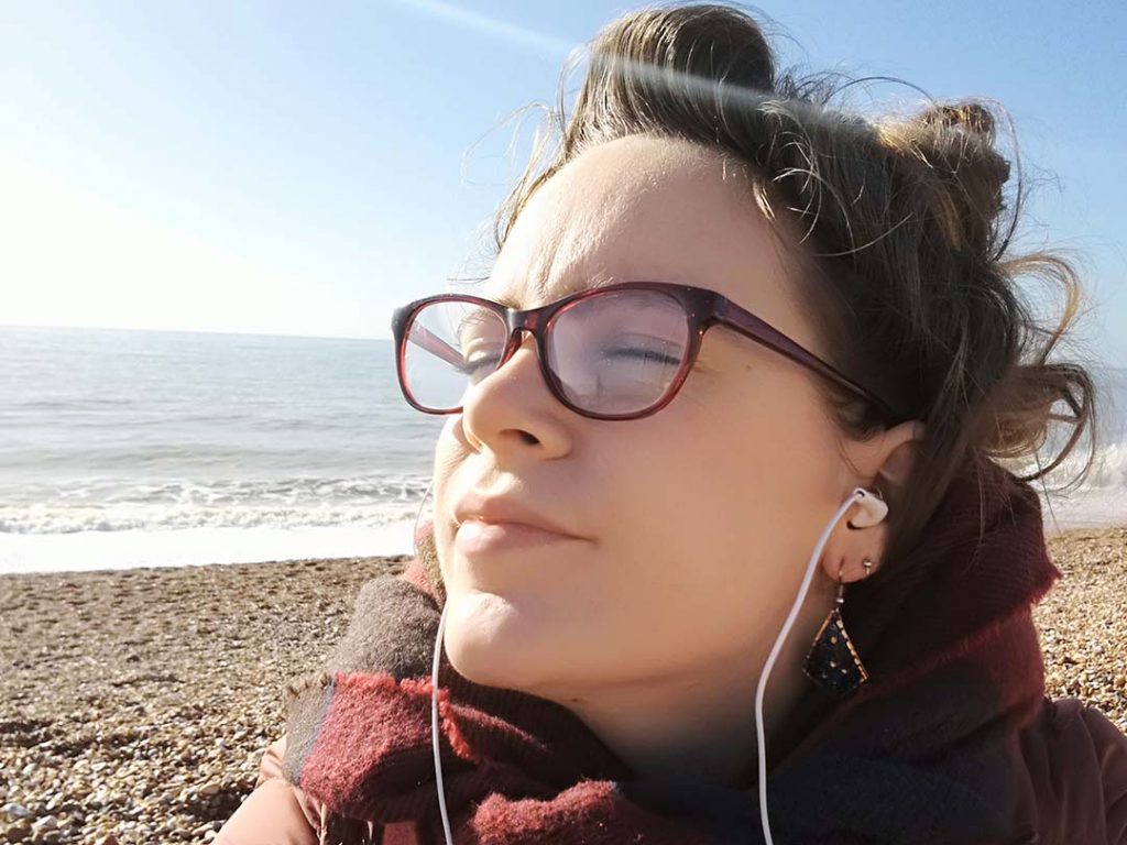 Alex is on a pebble beach with the sea in the background. She is wearing a pair of earphones.